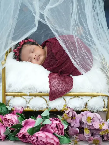 We ensure every newborn session is a comfortable and dreamy experience. Here, a peacefully sleeping baby is swaddled in a rich burgundy wrap, nestled in a miniature vintage-style bed. The soft fabrics, delicate floral accents, and gentle lighting all contribute to a serene and artistic infant portrait.