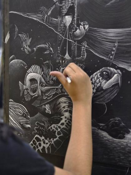 A close-up shot of an artist's hand adding intricate details to the fish and turtle in her surreal underwater scene using a glass marking pencil.