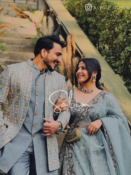 A happy couple during their celebrations. I designed the groom's outfit, a modern Indo-Western ensemble featuring a grey-blue kurta and a patterned open jacket, perfect for a sangeet or engagement ceremony.
