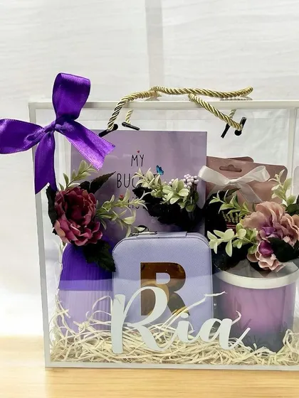 A close-up of the personalized acrylic gift bag for 'Ria'. The clear bag is tied with a purple ribbon and filled with items in a matching color palette, creating a cohesive and stylish look.