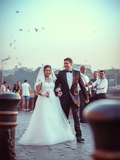 A candid shot of a couple in their wedding attire walking near the Gateway of India, with pigeons flying in the background.