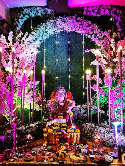 An elaborate and beautiful home decoration for the Ganpati festival. This setup features a floral arch with cherry blossom details, elegant candle-style lighting, and a green backdrop, creating a divine atmosphere to welcome Bappa into your home.