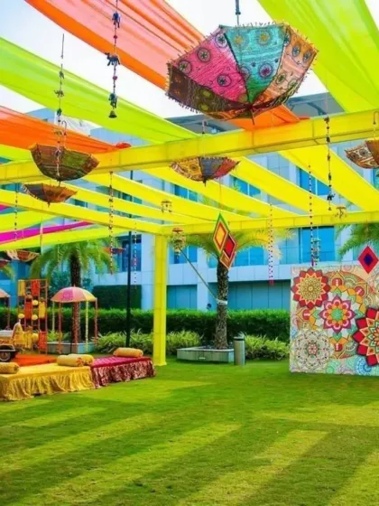 A spacious outdoor Mehendi setup on a lush green lawn. The decor features a canopy with hanging umbrellas and a colorful backdrop for photos.