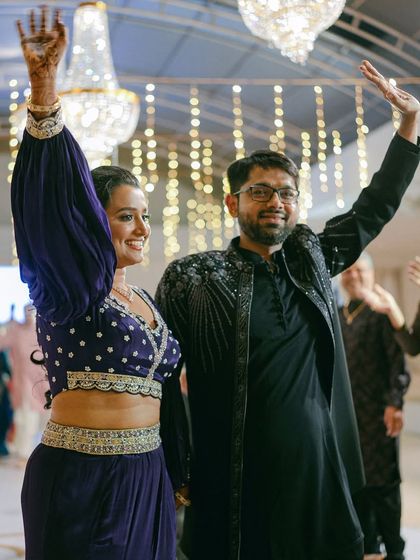 The bride and groom make a grand entrance, waving to their guests at the Sangeet. Their coordinated outfits in shades of blue and black match the event's decor.