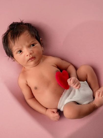 An awake newborn lies in a heart-shaped impression on a pink background, holding a small red heart prop.
