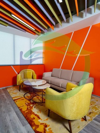This lounge area features large planters with Areca Palms and other foliage plants. The greenery softens the bold orange wall and complements the yellow and beige furniture, creating a balanced and inviting space for relaxation.