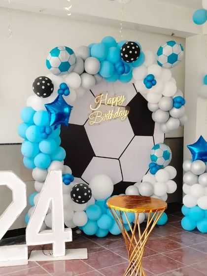 A football-themed birthday celebration for a 24th birthday. This setup features a large football-print backdrop, a blue, white, and silver balloon arch, and a light-up marquee number '24'.