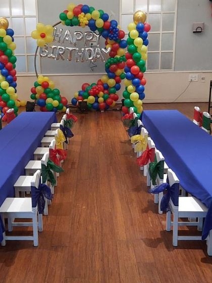 A colorful indoor party setup with long tables for kids and a heart-shaped balloon backdrop in primary colors.