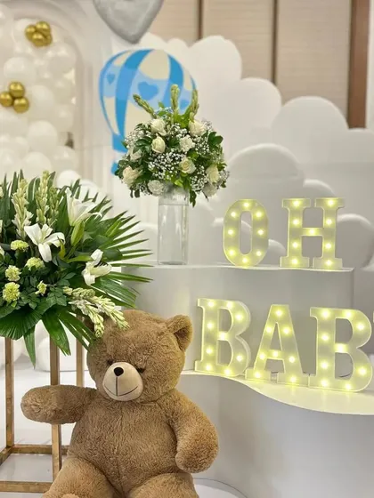 A close-up of the 'Oh Baby' marquee letters and teddy bear detail. The warm lights and lush floral arrangements add a touch of elegance and warmth to the overall setup.
