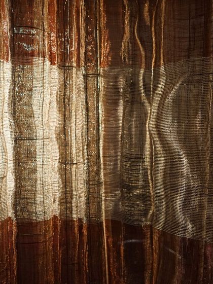 Light filters through a hand-woven panel from 'Cu+Fibers'. This close-up reveals the delicate structure of the weave, where copper and cotton yarns create an interplay of opacity and sheen.