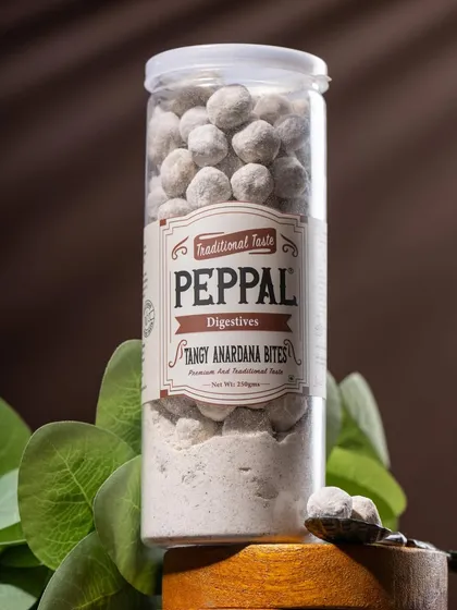 A clean product shot of Peppal's Tangy Anardana Bites. The styling with green leaves provides a fresh contrast to the earthy tones of the product and packaging.