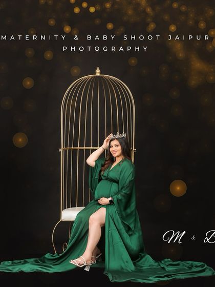 A regal studio portrait of a mom-to-be in a green gown and crown, seated in front of a golden birdcage. The dark, sparkly background creates a glamorous and enchanting mood.