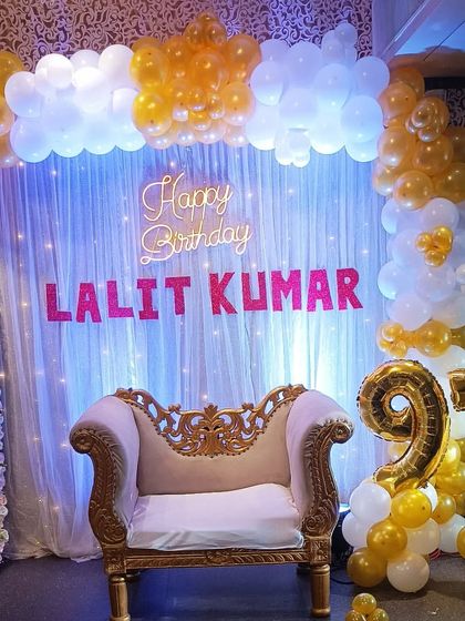 Celebrating a 97th birthday with a grand setup. This decoration includes a throne-style chair for the guest of honor, a backdrop of white curtains and fairy lights, and gold foil number balloons.