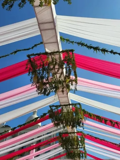 A view of the ceiling treatment for an outdoor Mehndi. Strips of pink and white fabric are stretched across the sky, with hanging floral chandeliers adding a touch of greenery and elegance.