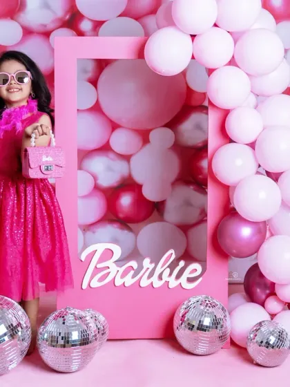 A vibrant 'Barbie' themed photoshoot for a stylish toddler. She poses confidently in a pink frame against a wall of pink balloons and disco balls, complete with a feather boa and sunglasses.