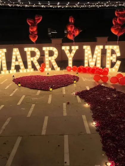This rooftop proposal setup makes a bold statement with giant 'MARRY ME' marquee letters. The romantic ambiance is enhanced by a heart made of red rose petals, candles, and overhead fairy lights.