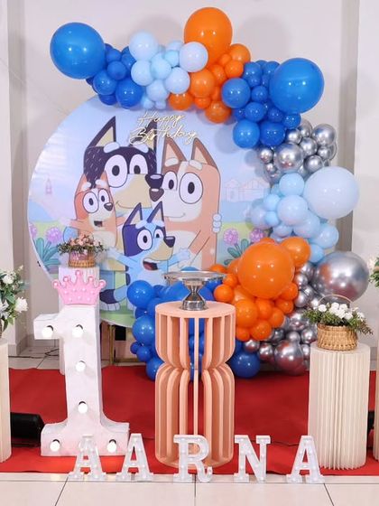 A charming Bluey themed first birthday party. The setup includes character cutouts of Bluey and Bingo, a custom backdrop, and a blue and orange balloon arrangement.