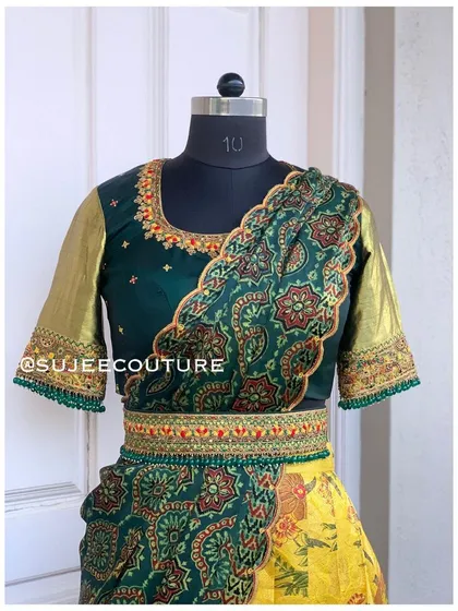 A unique half saree design featuring a yellow Kalamkari print skirt and a dark green blouse. The look is tied together with a matching embroidered vaddanam and a draped green dupatta.