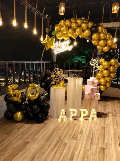 A stylish 50th birthday party for "Appa" on a terrace. The setup features a black and gold balloon ring, a personalized light-up name sign, and foil balloons marking the golden jubilee.