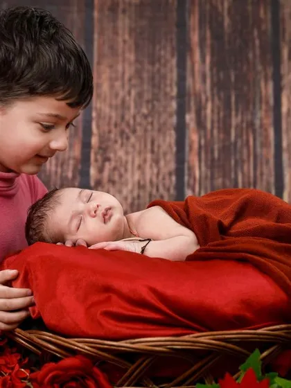 An older brother looks lovingly at his new baby sibling, who is nestled in a basket with a rich red blanket.