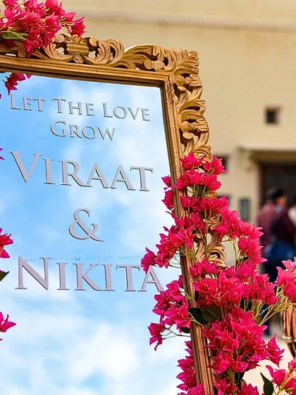 A creative welcome sign for a Mehendi, with the couple's names etched onto a mirror and framed with vibrant bougainvillea. The reflection of the sky adds a beautiful, dynamic element.