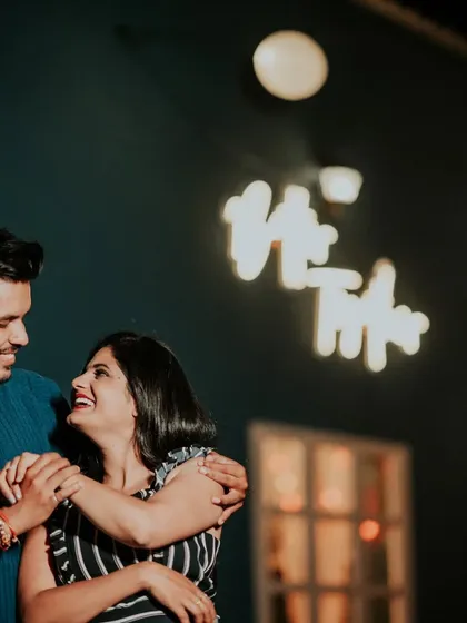 A candid, happy moment at a cafe-style night setup. The warm glow of the neon sign and string lights creates a cozy and intimate atmosphere.