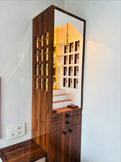 A full view of a custom wooden mandir with intricate side panels featuring bell cutouts. The unit includes drawers and cabinets at the base for storing puja items.