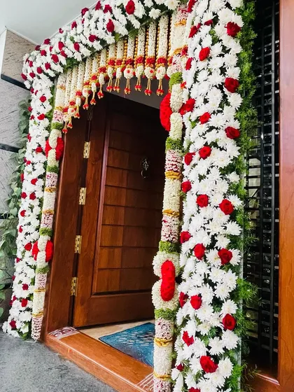 A close-up of a classic floral door decoration. The frame is covered in white and red flowers, with traditional hanging elements that add a touch of grace to the home's entrance.