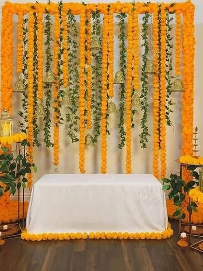 I created this indoor Haldi backdrop for a traditional home ceremony. It uses dense strings of bright orange marigolds accented with green leaves and hanging temple bells. The setup includes a clean, white-covered seat and matching floral arrangements on stands for a complete and authentic look.