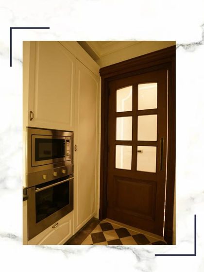 A glimpse into the kitchen from the doorway, showing the built-in oven and microwave stack, classic white cabinets, and a beautiful glass-paned wooden door.