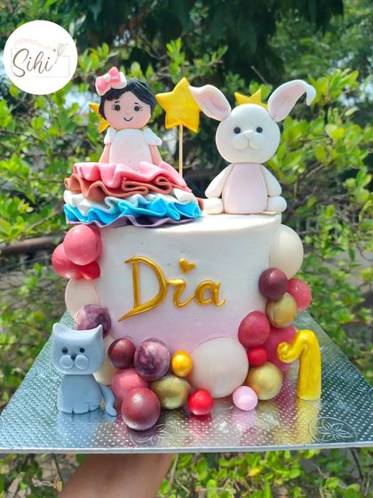 A colorful and cute first birthday cake. It features a custom girl figurine alongside her favorite bunny and cat friends, surrounded by a cascade of multi-colored baubles.