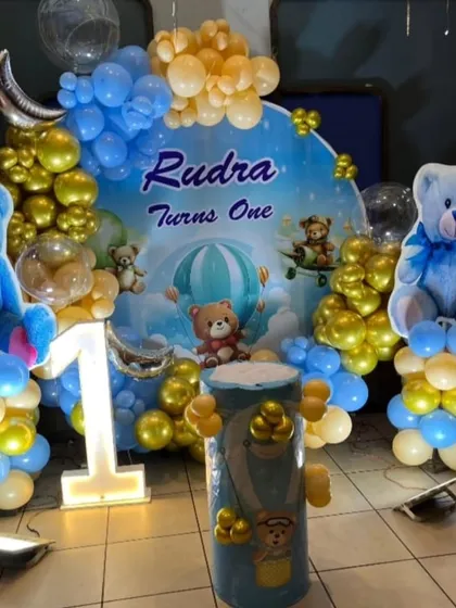 A detailed look at the adorable teddy bear themed first birthday backdrop. The design features bears floating in hot air balloons against a sky-blue background, framed by blue and gold balloons.