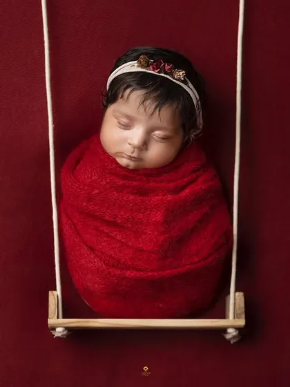 A simple and creative shot of a baby swaddled in a bold red wrap, sleeping on a tiny swing. The solid red background makes the image pop.