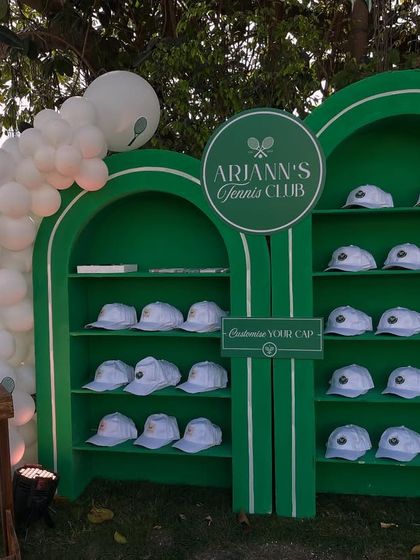 A custom 'Tennis Club' favour station where each guest received a personalized cap. The green shelving unit is decorated with a white balloon garland.
