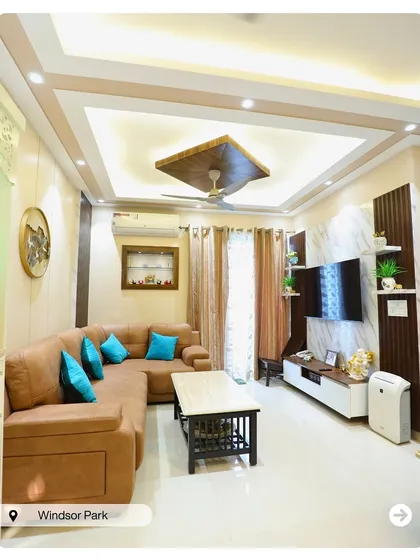 This living room at Windsor Park features a comfortable brown sectional sofa with bright blue cushions for a pop of color. The false ceiling design with a central wooden element adds a unique touch.