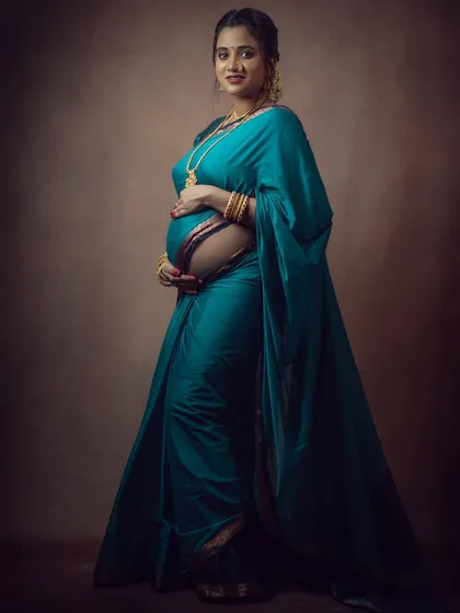 A beautiful studio maternity portrait of a woman in a teal saree, looking at the camera with a gentle smile.