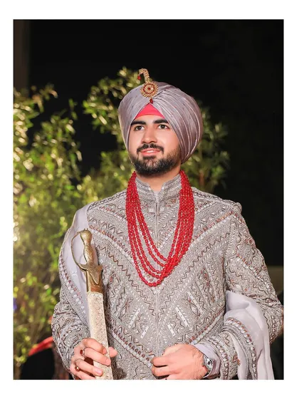 A classic portrait of a Sikh groom in his grey embroidered sherwani and red beaded necklace. His confident stance and the traditional kirpan create a powerful image.