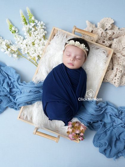 A serene composition in shades of blue. The baby is swaddled in navy and resting in a wooden tray, surrounded by flowing fabrics and delicate flowers.