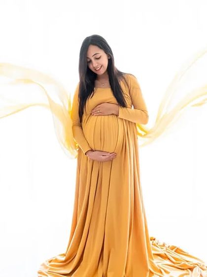 A graceful solo portrait featuring a flowing yellow maternity gown against a bright white backdrop. The soft fabric toss adds a touch of dynamic elegance to the shot.