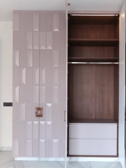 Inside, the wardrobe features a rich wood-grain laminate, providing a warm contrast to the exterior. The layout includes hanging space and deep drawers for versatile storage.