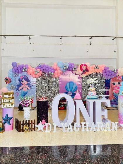 A full stage view of a mermaid-themed first birthday. The setup includes a silver shimmer wall, character cutouts, 'ONE' marquee letters, and an underwater-themed balloon arch.