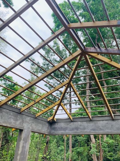 The exposed roof structure of our ongoing Coorg project. The wooden framework, set against the backdrop of the forest canopy, shows the bones of the building before the terracotta tiles are laid, highlighting the craftsmanship involved.