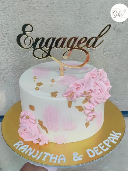 A simple and sweet pink and white engagement cake. The textured buttercream is accented with gold flecks and piped pink rosettes, topped with an elegant "Engaged" acrylic topper.