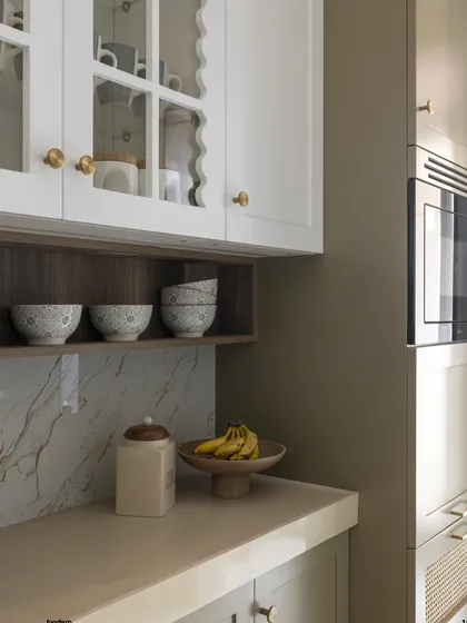 A quiet corner of a kitchen, showcasing how different elements come together. The scalloped edge of the white cabinet, the warm wood of the open shelf, and the subtle veining of the marble backsplash create a soft and inviting composition.