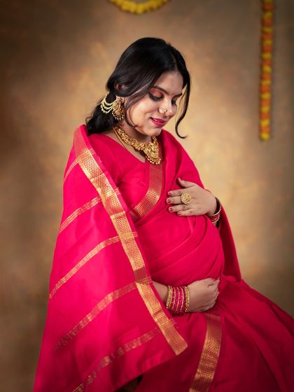 A close-up portrait capturing the gentle emotion of an expecting mother in a red saree. Her traditional gold jewelry and the soft lighting highlight the beauty and heroism of motherhood.