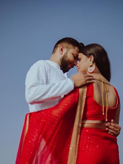 A minimalist and powerful couple portrait against a clear blue sky. The simplicity of the background puts all the focus on their embrace and the striking contrast of the bride's red saree.