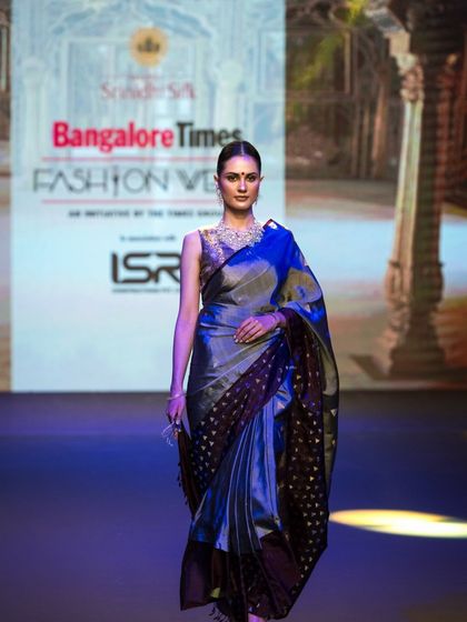 A full-length runway shot of a model in a shimmering blue and black saree. The lighting highlights the fabric's texture and sheen, an important detail for designers and buyers.