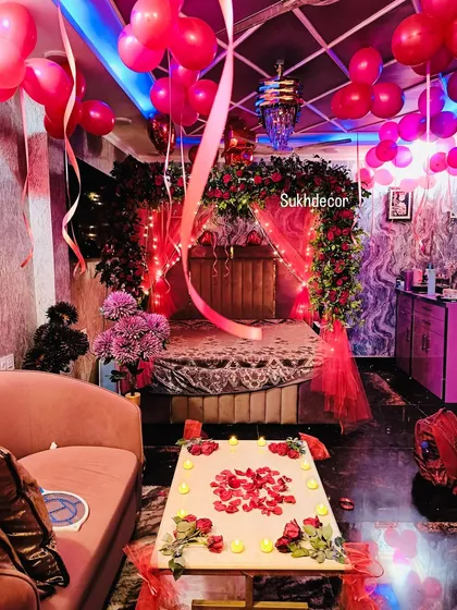 I design intimate and romantic room decorations perfect for a pre-wedding surprise. This setup includes a bed canopy made of red roses and greenery, fairy lights, red balloons on the ceiling, and a candlelit table with rose petals.