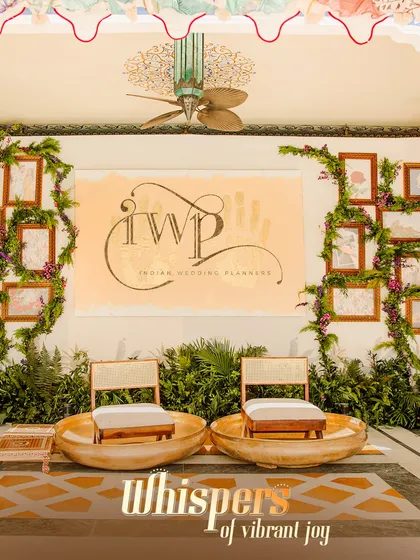 A personalized Mehendi seating area with our IWP logo and handprint art in the background. The seating is adorned with fresh greenery, making it a beautiful focal point for the bride.