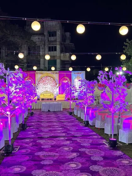 The full view of an outdoor wedding reception area with a purple and pink theme. The long aisle is lined with illuminated decorative trees, leading the eye directly to the grand stage.